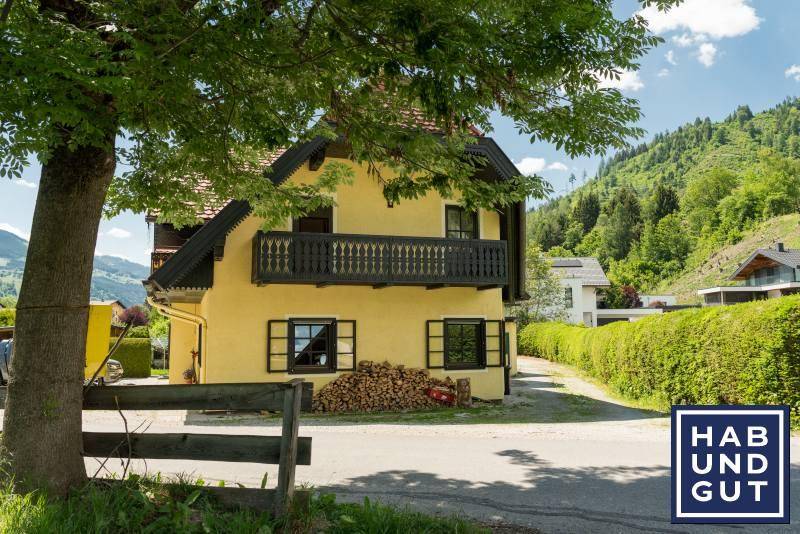 Gelbes Haus mit Holzbalkon und rotem Dach, umgeben von Bäumen und Bergen.