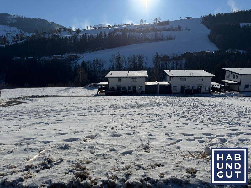 Schneebedeckte Landschaft mit Häusern und einer Skipiste im Hintergrund an einem sonnigen Wintertag.