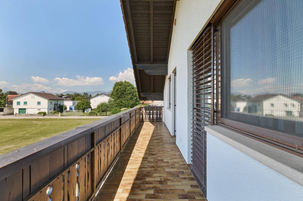 Langer Balkon mit Holzboden und Blick auf die grüne Umgebung und Nachbarhäuser.
