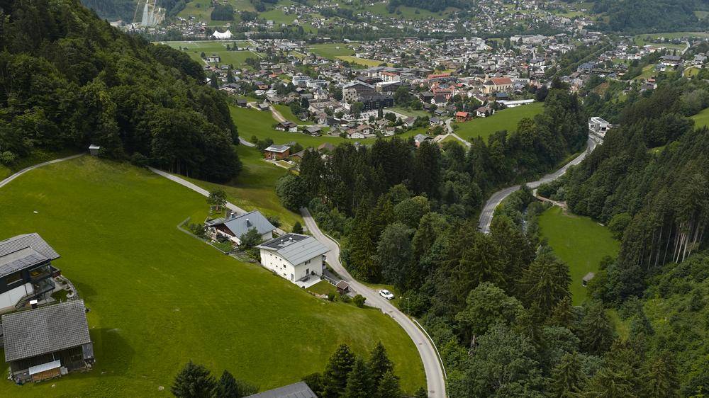 Luftaufnahme einer kurvenreichen Straße durch ein grünes Tal mit Häusern und einer Stadt in der Ferne.
