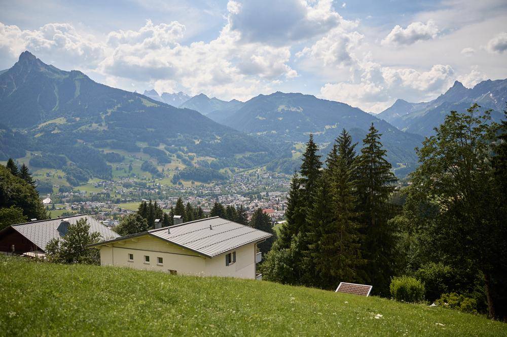 Haus auf einem grünen Hang mit Blick auf eine Stadt im Tal und majestätische Berge unter bewölktem Himmel.