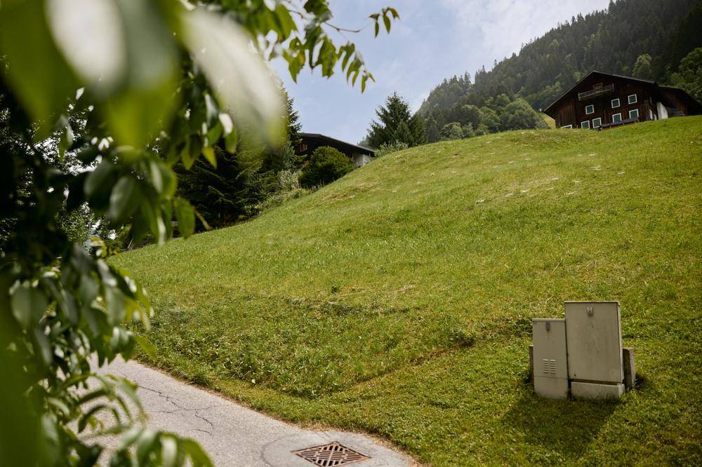 Grüne Hanglandschaft mit einem kleinen Weg und Häusern im Hintergrund, daneben stehen Versorgungskästen.