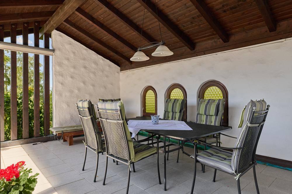 Überdachte Loggia mit Esstisch, Stühlen und rustikalen Rundbogenfenstern.
