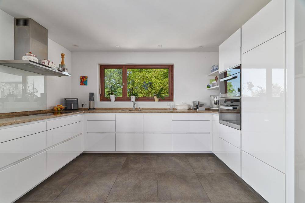 Helle, moderne Küche mit weißen Hochglanzfronten, integrierten Geräten und Fensterblick ins Grüne.