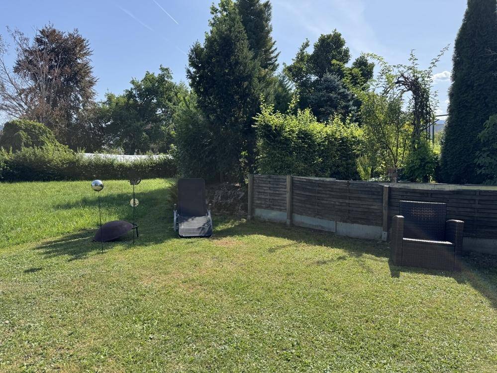 Gepflegter Gartenbereich mit Rasenfläche, Sitzgelegenheiten und üppiger Bepflanzung unter blauem Himmel.
