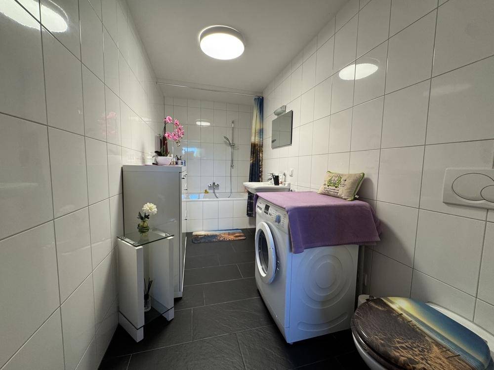 Funktionales Badezimmer mit weißen Wandfliesen, dunklem Boden, Badewanne mit Duschfunktion und Waschmaschine.