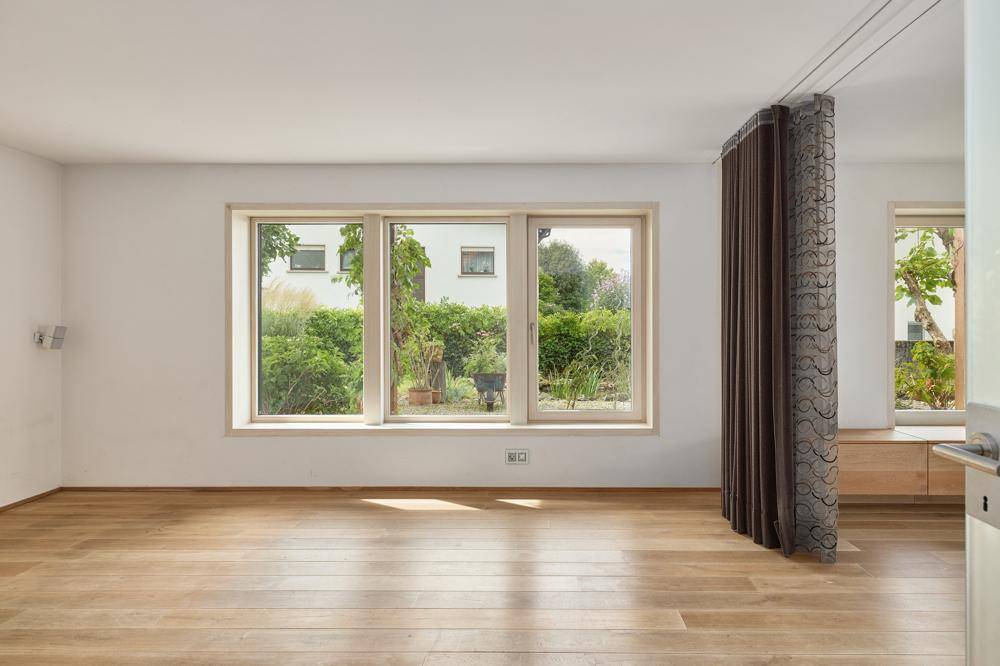 Heller, leerer Raum mit großen Fenstern und Blick ins Grüne, ausgestattet mit Holzfußboden.