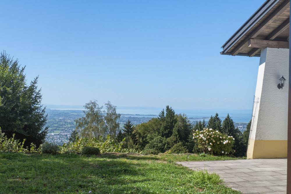 Teilansicht des Hauses mit Terrasse und Rasenfläche, umgeben von Bäumen und mit Seeblick.