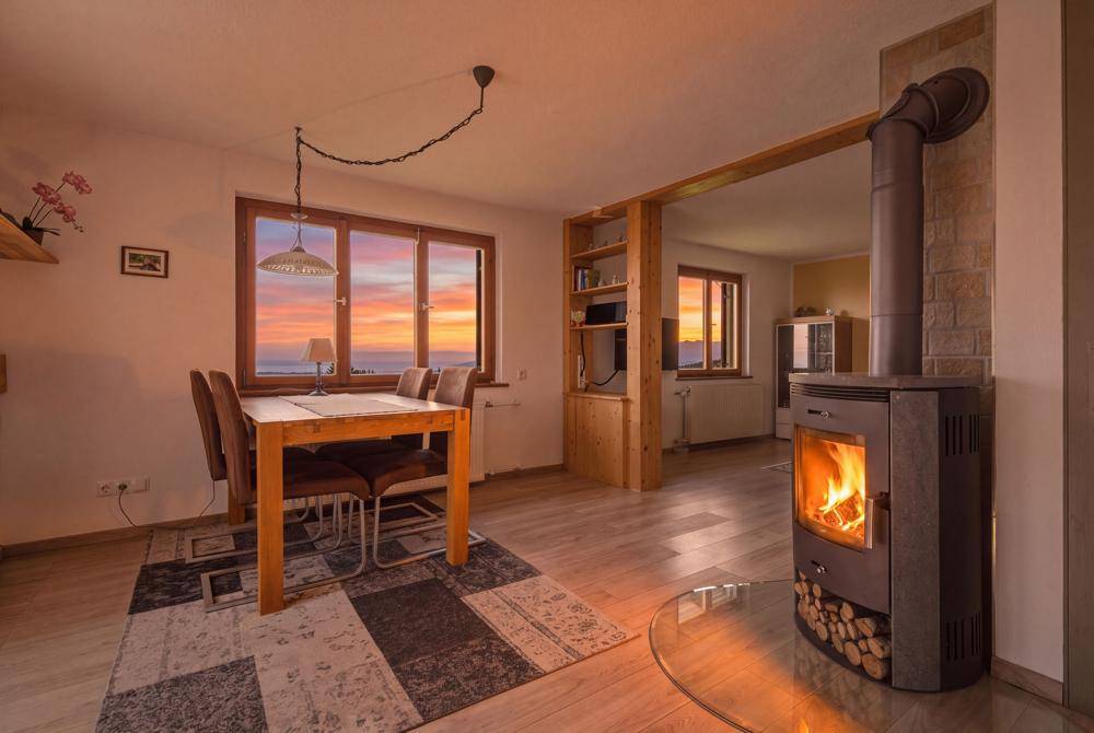 Wohn-Essbereich mit Kaminofen und Holzboden, beleuchtet vom warmen Licht des Sonnenuntergangs.