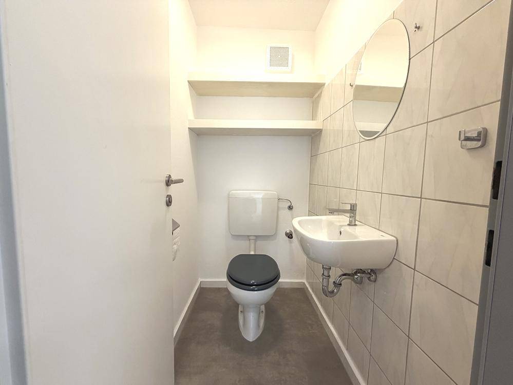 Kleines Gäste-WC mit Toilette, Waschbecken und rundem Spiegel, funktional und hell gefliest.