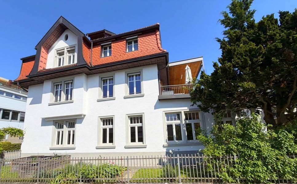 Weißes Gebäude mit rotem Mansarddach und Balkon unter blauem Himmel.