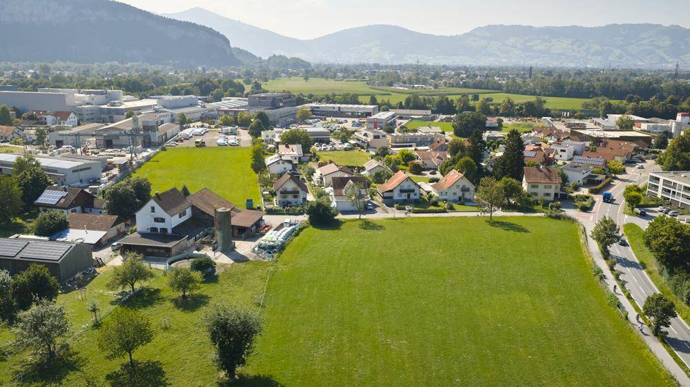 Weitläufige Luftaufnahme einer ländlichen Siedlung mit grünen Feldern und Bergen im Hintergrund.