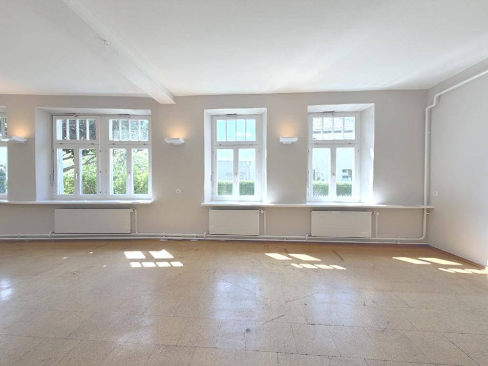 Heller, leerer Raum mit vielen Fenstern und Holzboden.