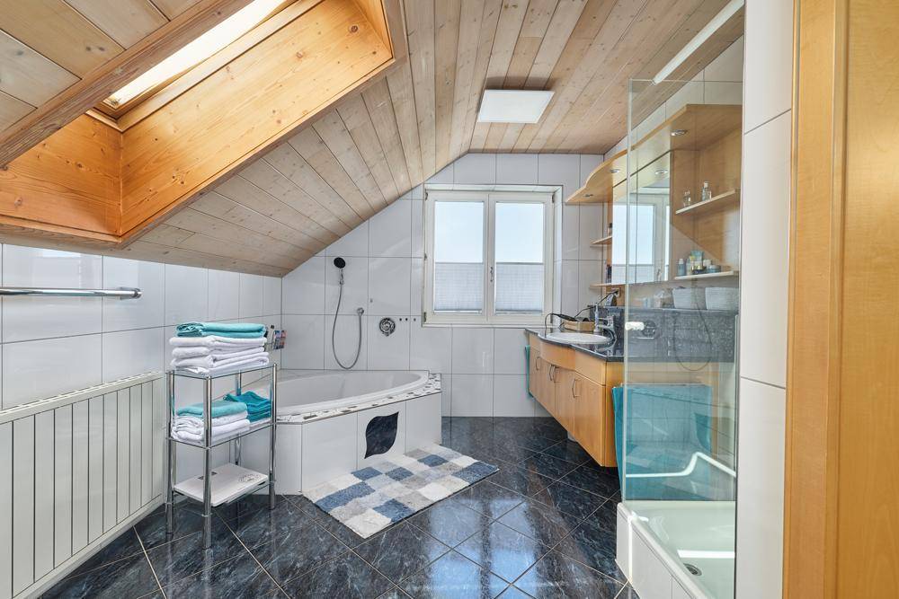 Badezimmer im Dachgeschoss mit Badewanne, Dusche, Dachfenster und dunklen Fliesen.