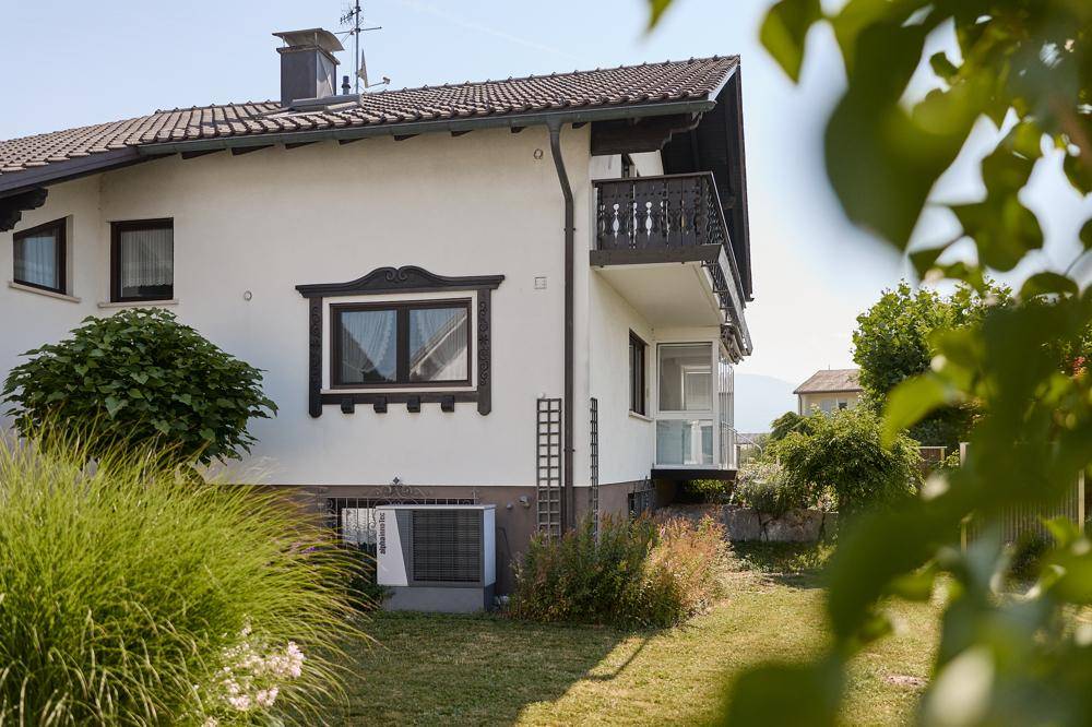 Weißes Einfamilienhaus mit Satteldach, Balkon und einer Wärmepumpe im Vorgartenbereich.