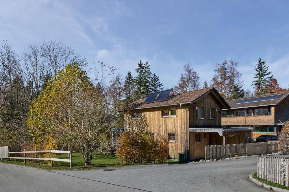 Holzhaus in ländlicher Umgebung mit Zaun und Bäumen unter blauem Himmel.