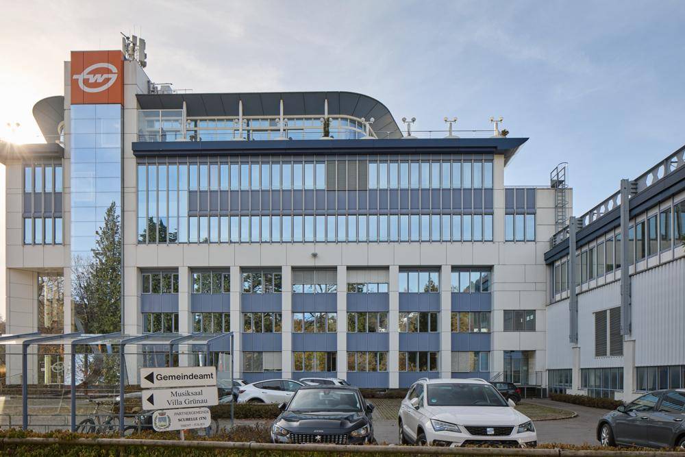 Moderne Bürogebäude-Fassade mit großen Glasflächen und einem Parkplatz davor unter hellem Himmel.
