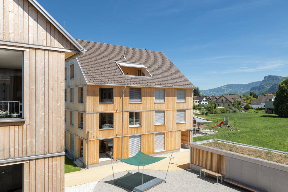 Modernes Holzgebäude mit einem gemeinschaftlichen Innenhof und Spielbereich vor bergiger Kulisse.