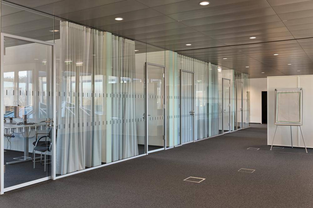 Moderner Bürogang mit Glaswänden, Teppichboden und Türen zu einzelnen Büros.