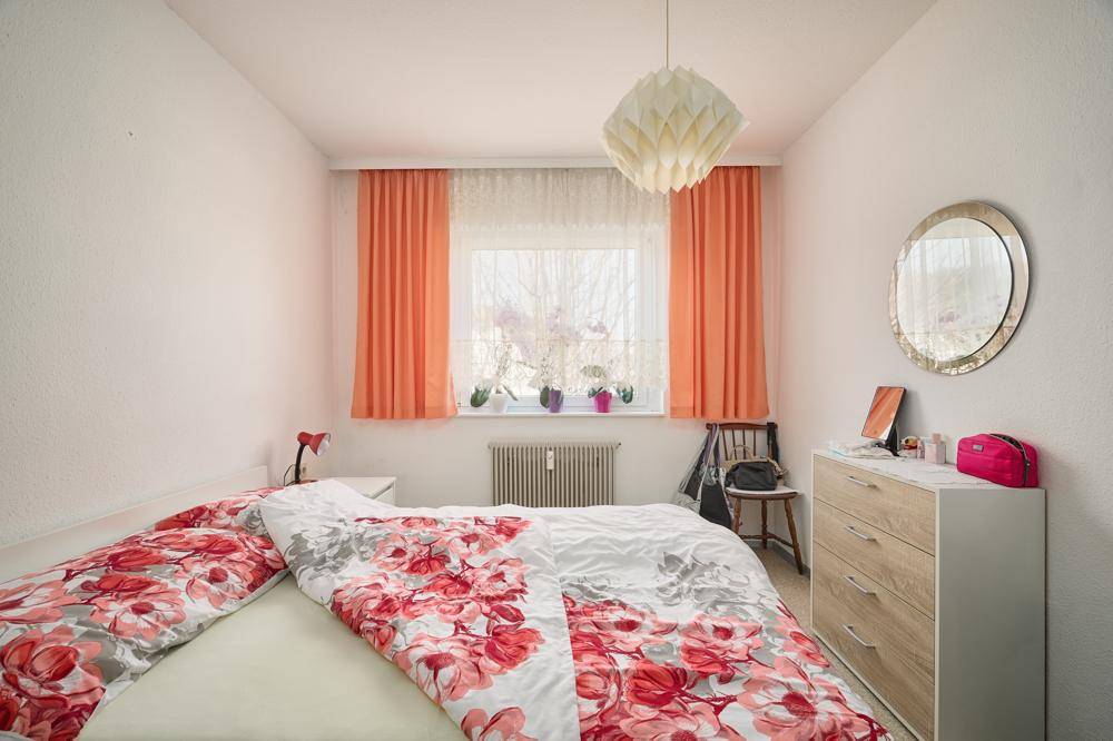 Schlafzimmer mit Doppelbett, Kommode, Spiegel und Fenster mit orangefarbenen Vorhängen.