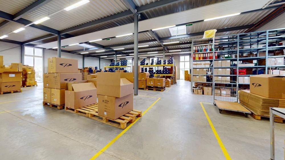 Innenansicht der Lagerhalle mit gestapelten Kartons, einem Rollwagen und umfangreichen Regalsystemen.