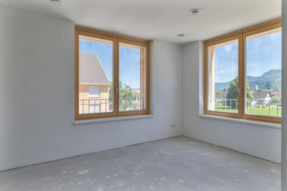 Heller, unmöblierter Raum mit großen Holzfenstern und Blick auf die umliegende Landschaft und Berge.