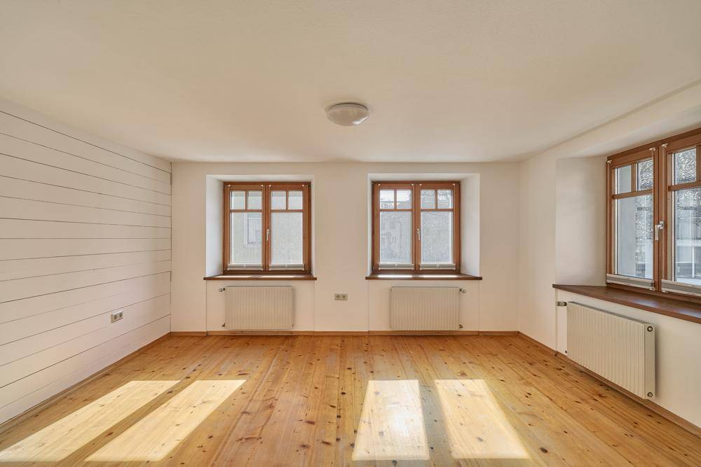 Leerer Raum mit hellem Holzfußboden und mehreren Fenstern, gut belichtet.
