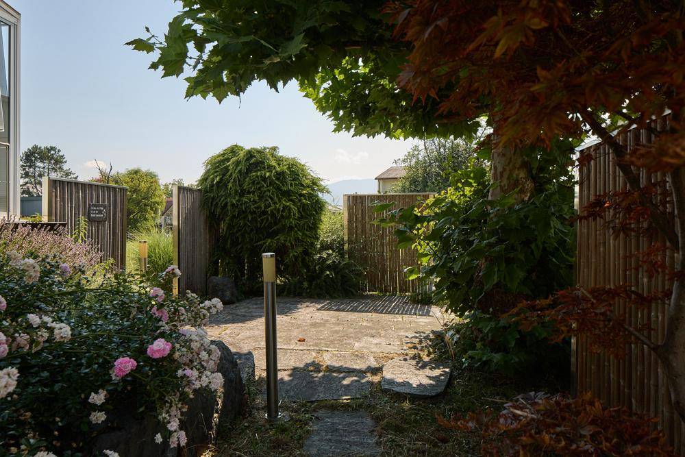 Gepflegter Gartenbereich mit Steinplatten, Bambuszaun und blühenden Sträuchern unter Bäumen.