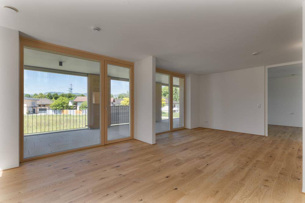 Heller, leerer Raum mit Holzboden und großen Fenstern, die auf einen Balkon führen.