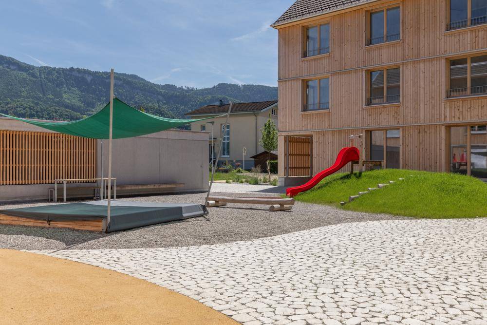 Detailansicht eines Spielplatzes mit roter Rutsche und Sonnensegel vor der modernen Holzfassade.