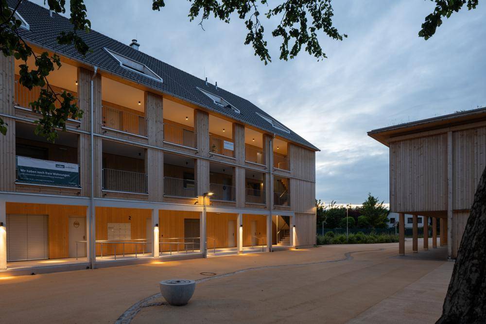 Fassade eines modernen Gebäudes mit Holzverkleidung und beleuchteten Balkonen in der Abenddämmerung.