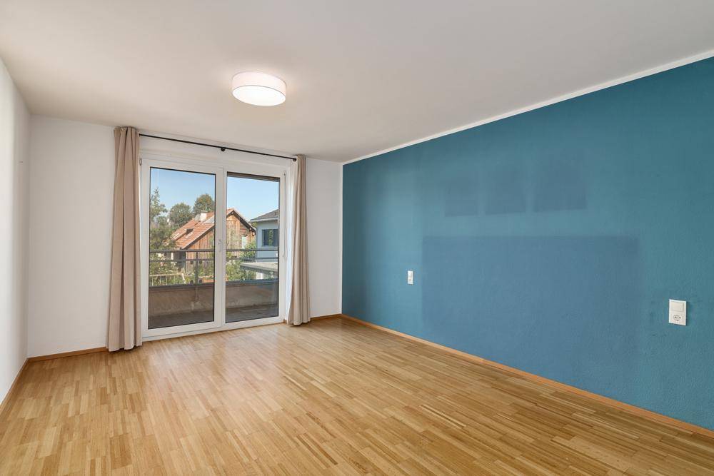 Helles Zimmer mit Holzboden, blauer Akzentwand und Balkonzugang.