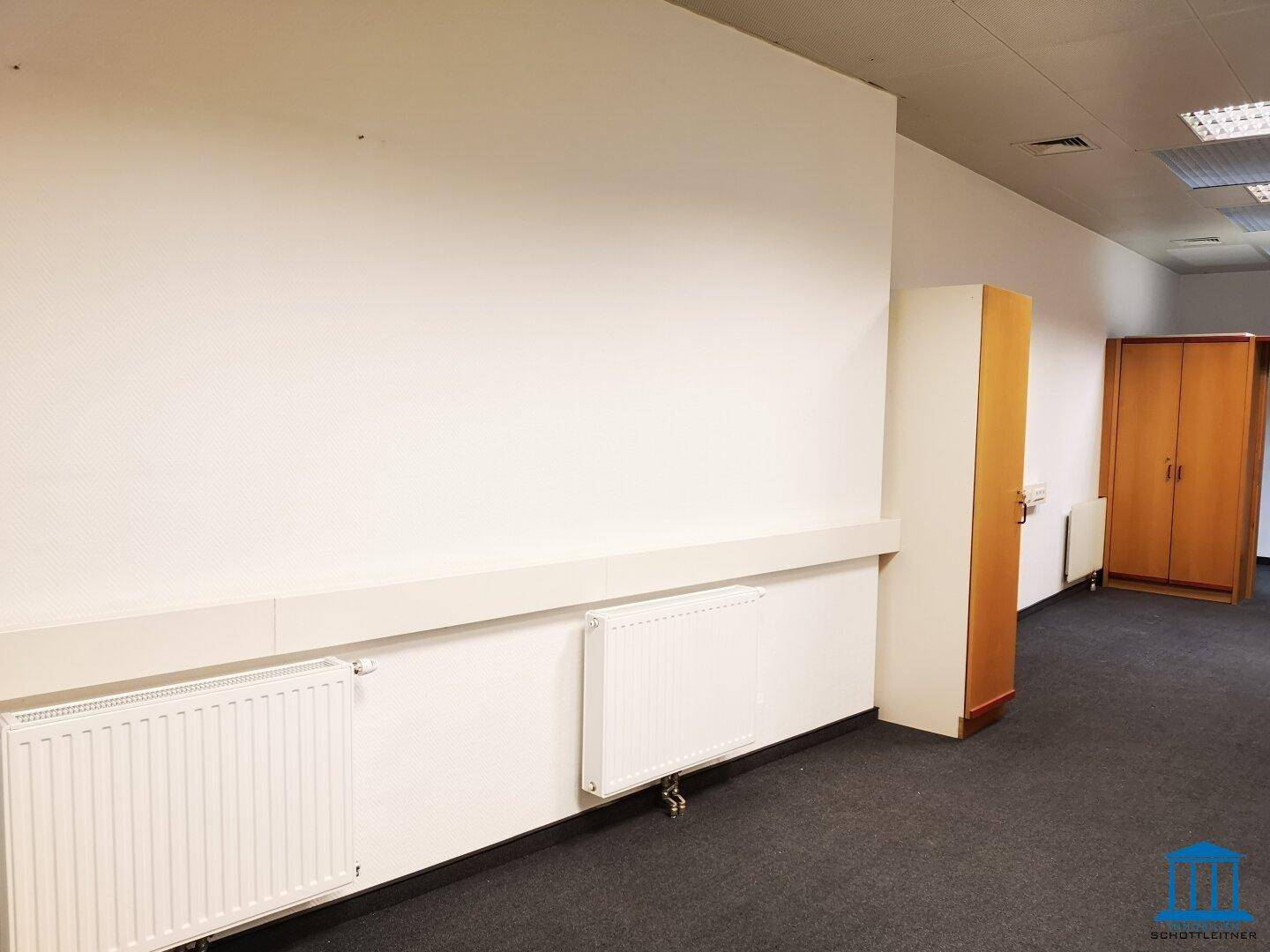 Langer Bürobereich mit Teppichboden, Heizkörpern und einem Schrank, ideal für Großraumbüro oder Flur.