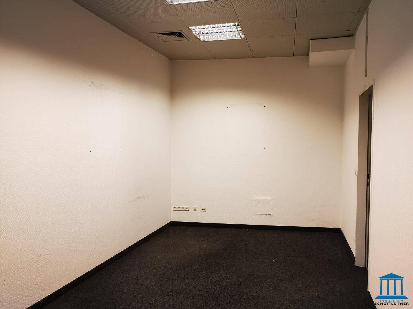 Leerer Büroraum mit Teppichboden und Steckdosen an der Wand, bereit für neue Mieter.