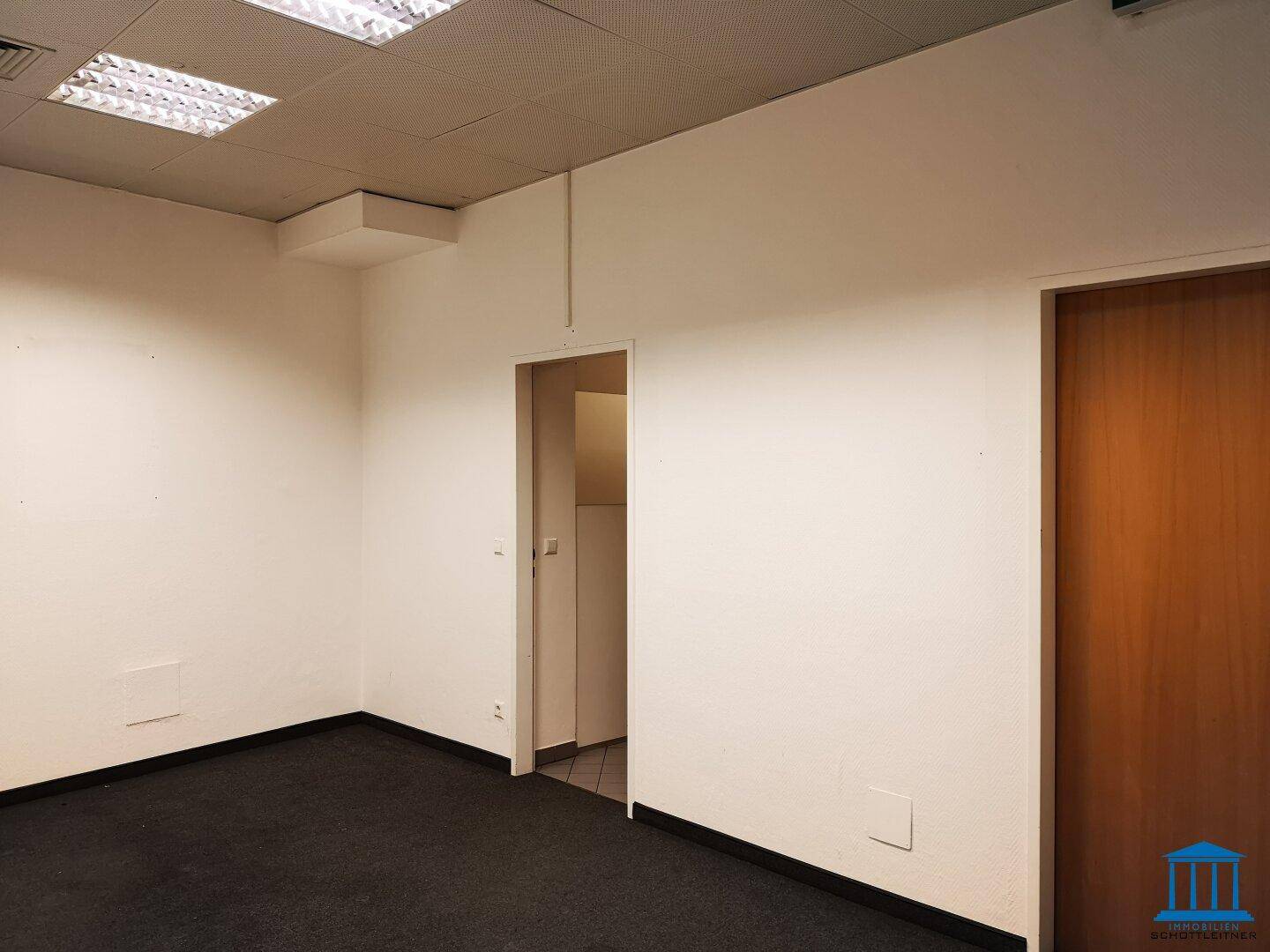 Heller Büroraum mit Teppichboden und einer Holztür, ideal für ein Einzelbüro oder Besprechungszimmer.