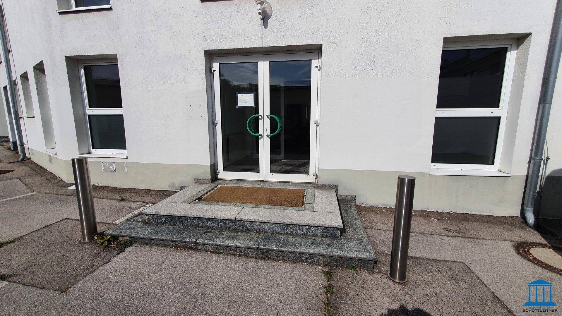 Haupteingang eines Gewerbegebäudes mit Glastüren und Granitstufen, umgeben von Parkplätzen.