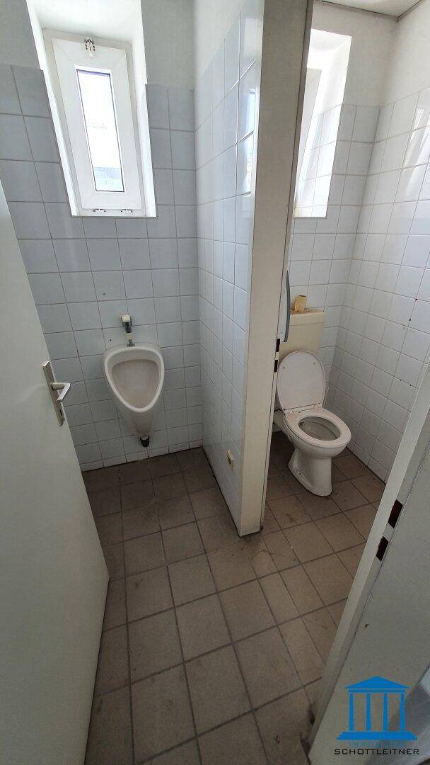 Gefliestes WC mit Urinal und Toilette, durch eine Trennwand separiert, mit kleinem Fenster.