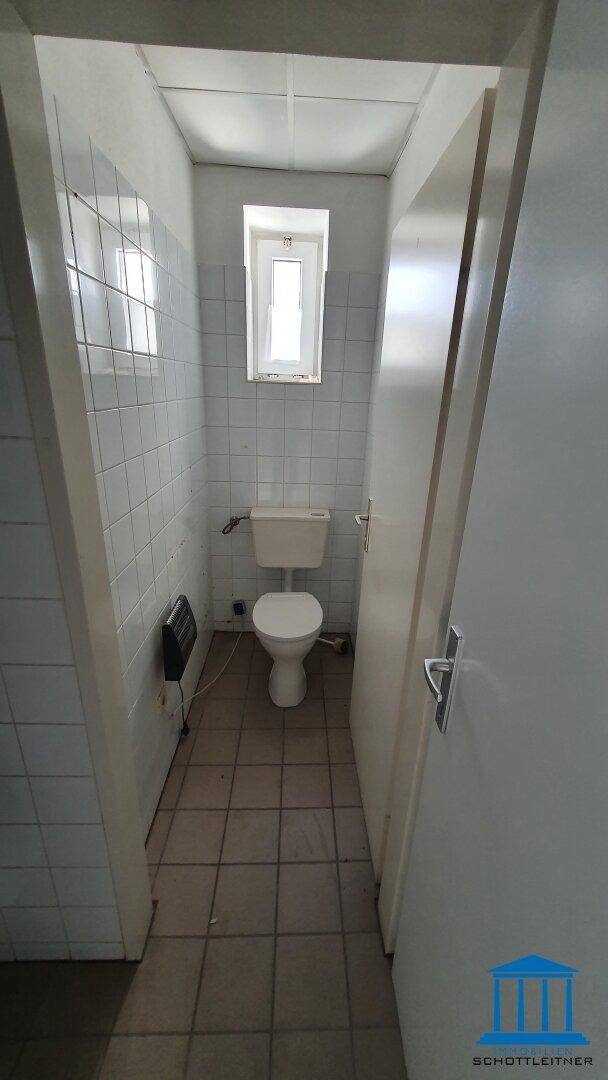 Schmale Toilettenkabine mit WC, gefliesten Wänden und einem kleinen Fenster für Tageslicht.