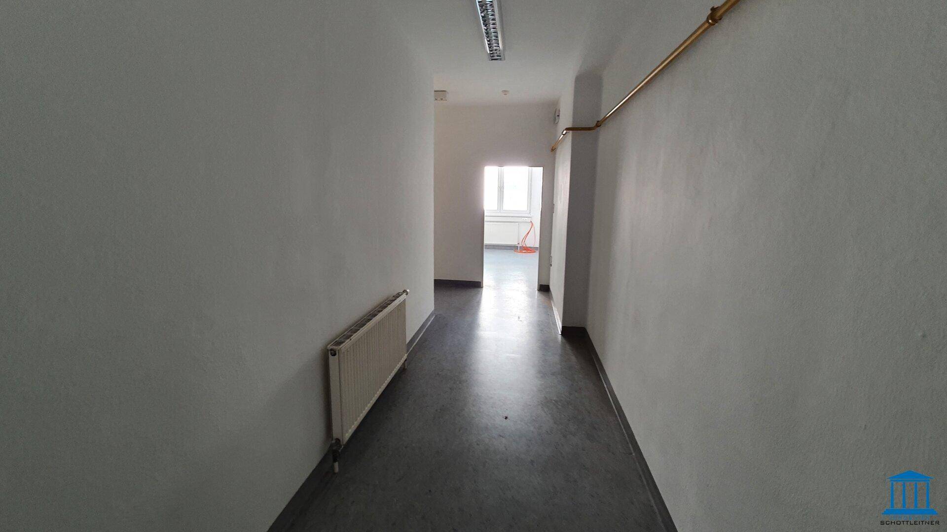 Schmaler Flur mit Heizkörper und Blick in einen weiteren Raum, ausgestattet mit Linoleumboden.