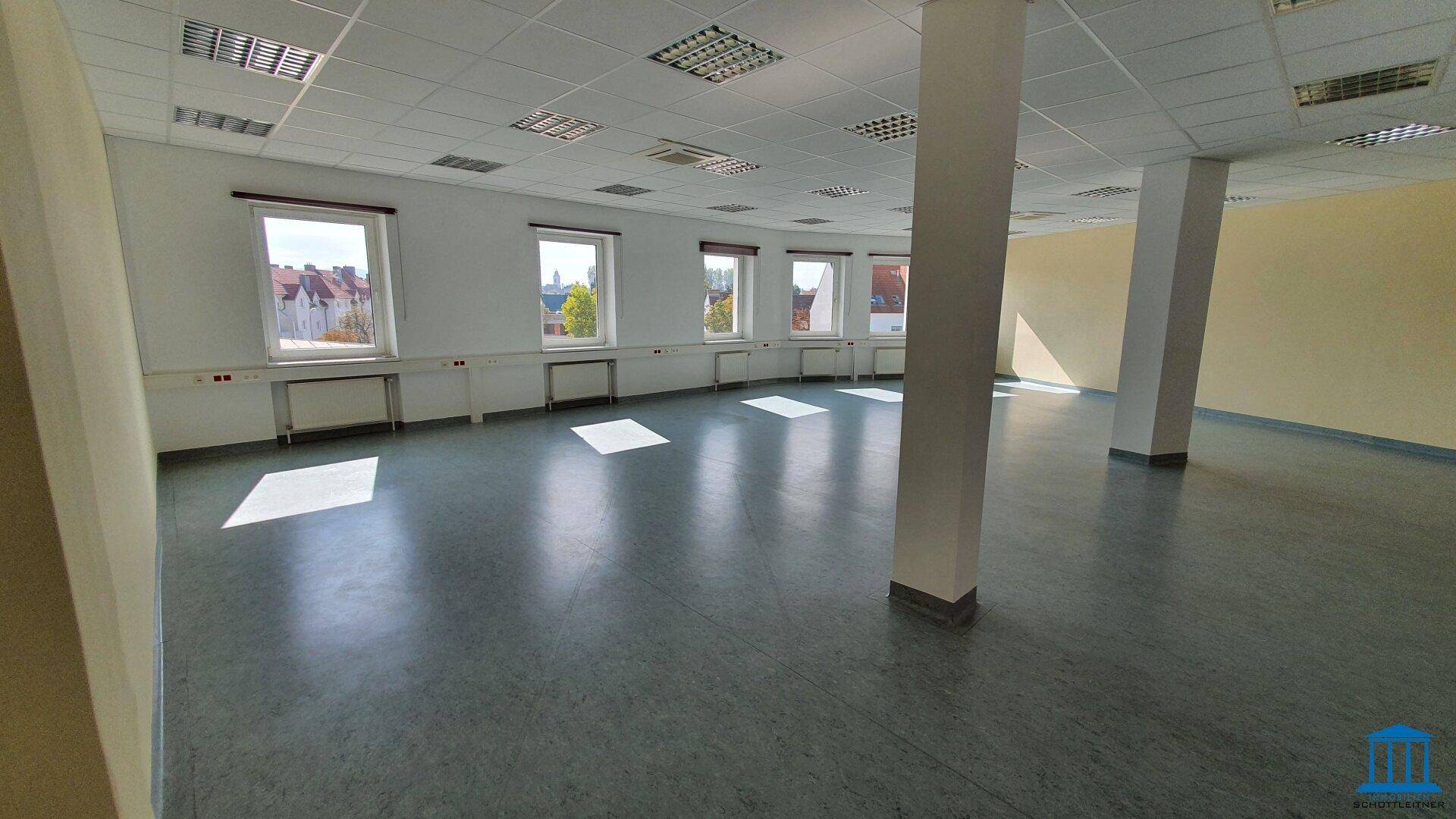 Heller Büroraum mit vielen Fenstern entlang einer gekrümmten Wand und Säulen, ausgestattet mit Linoleumboden.