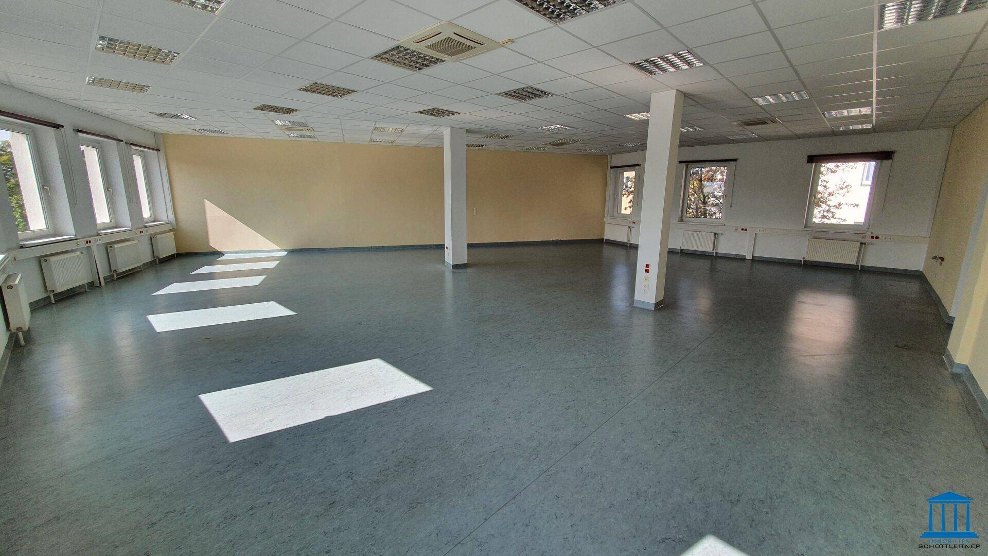 Geräumiger Bürobereich mit mehreren Fenstern, Säulen und grauem Bodenbelag, gut beleuchtet durch Tageslicht.