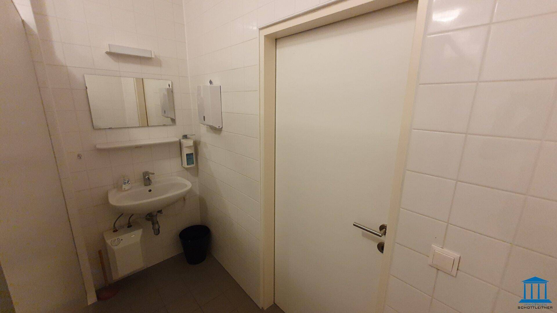 Badezimmer mit Waschbecken, Spiegel und Seifenspender, die Tür führt zu einer weiteren Kabine.