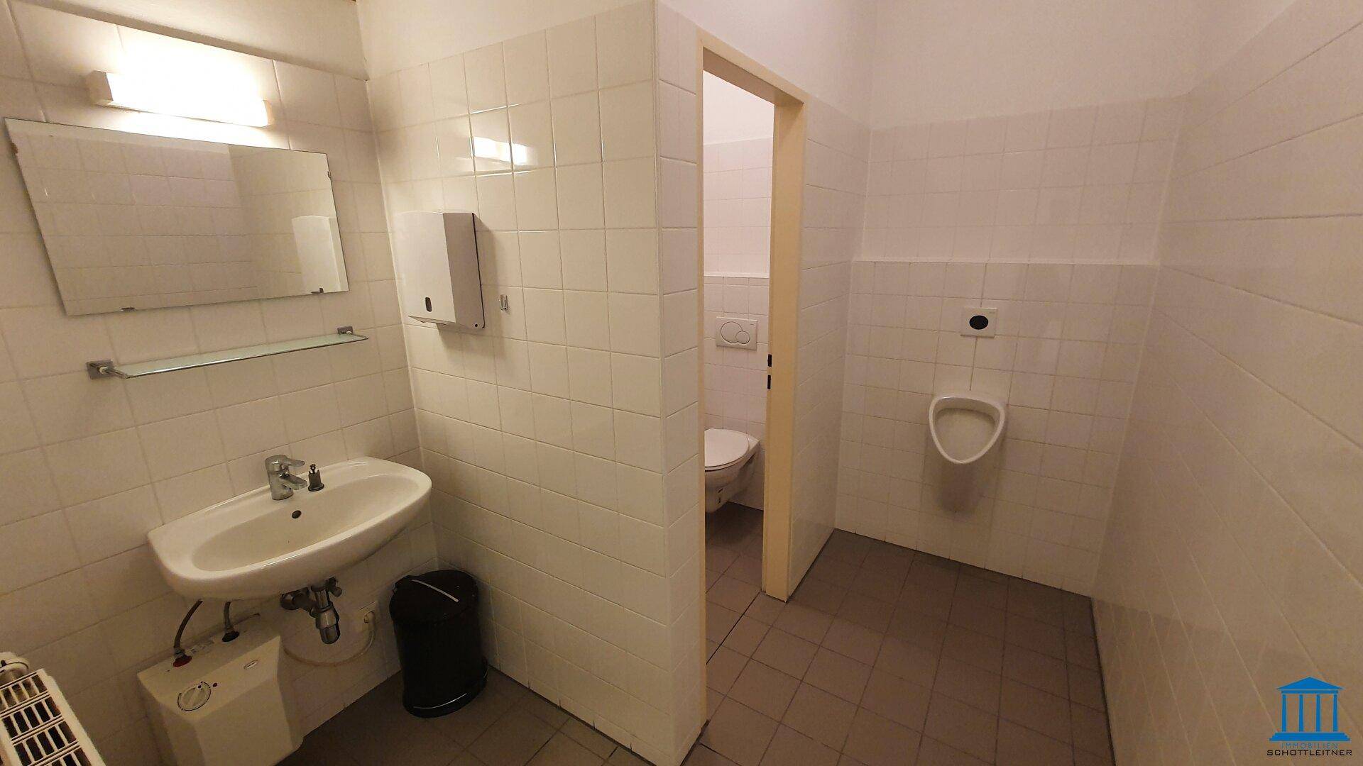 Badezimmer mit Waschbecken, Spiegel, Urinal und Zugang zu einer Toilettenkabine.
