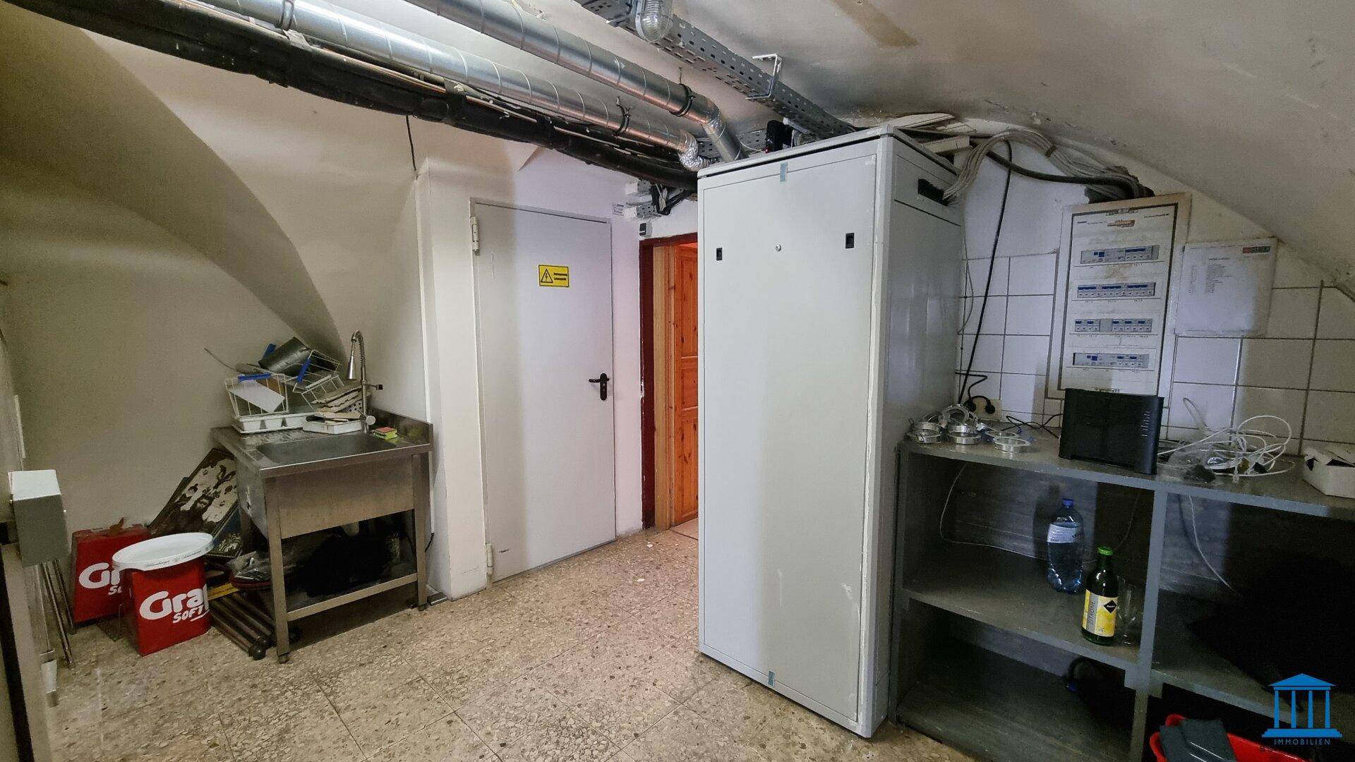 Lagerraum mit Regalen, Spüle und technischen Installationen an der Decke.