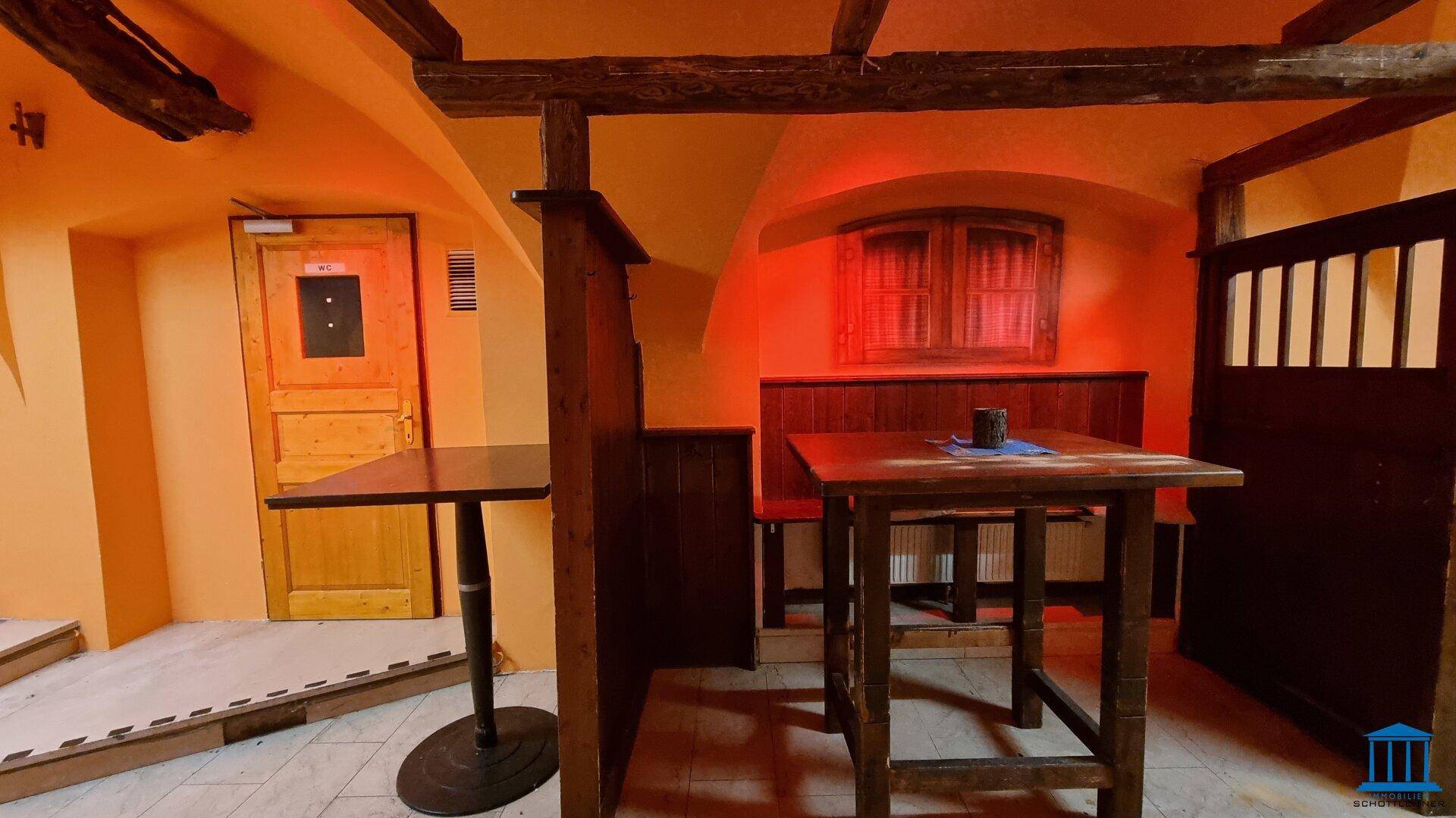 Rustikaler Innenbereich mit Holzbalken, Tischen und Bänken, beleuchtet in warmem Orange.
