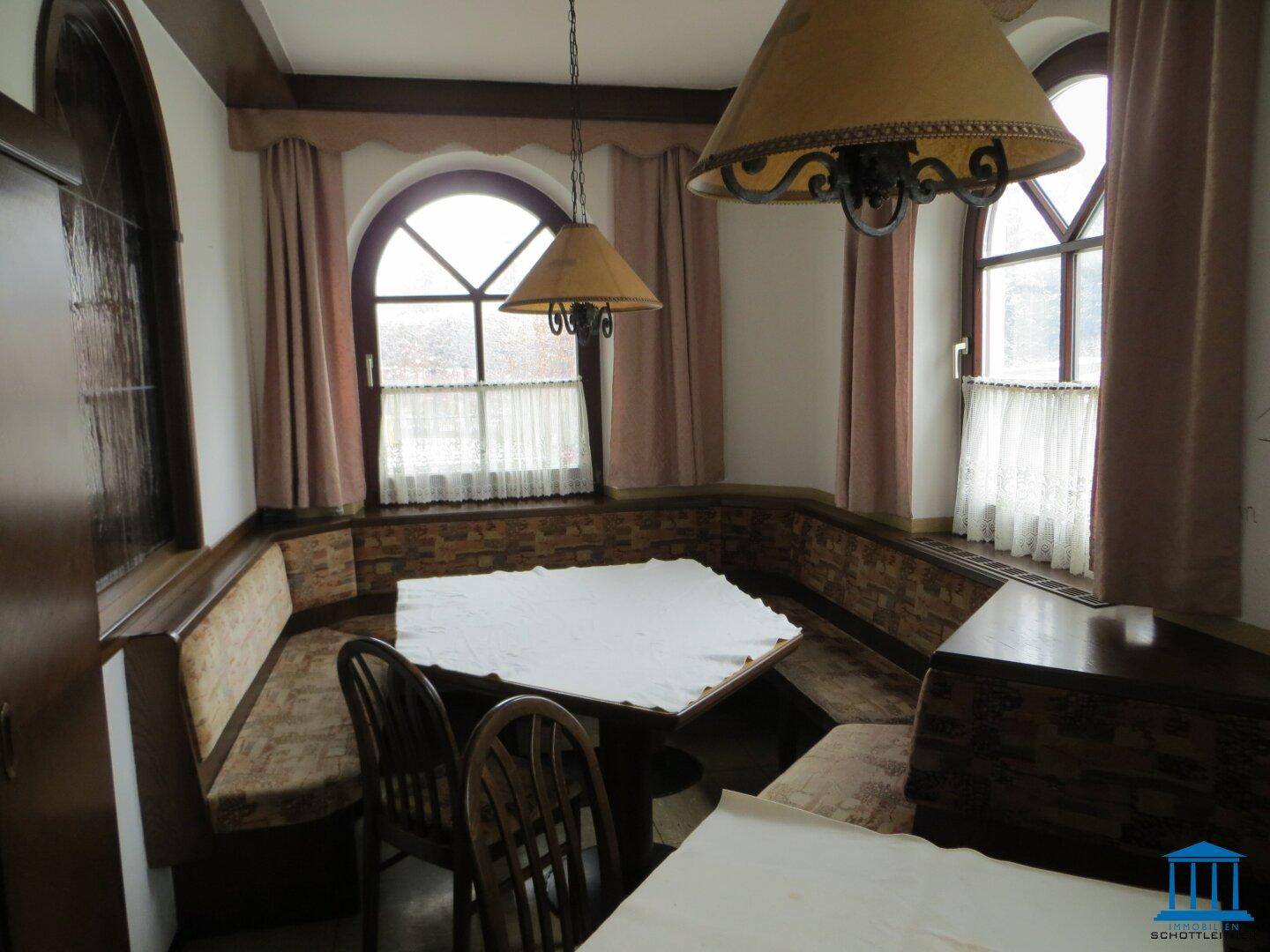 Eckbankbereich in einem traditionellen Speisesaal mit Holztischen und Rundbogenfenstern.