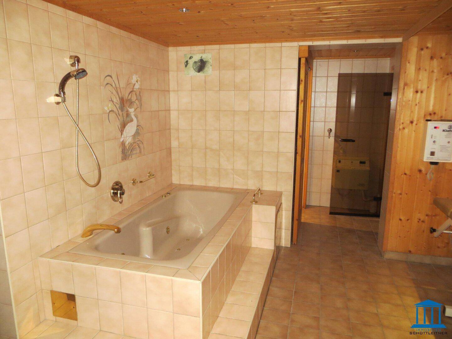 Gefliestes Badezimmer mit eingebauter Badewanne, Duschvorrichtung und Zugang zu einer Sauna.
