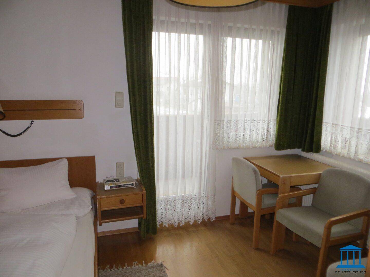 Helles Schlafzimmer mit Einzelbett, Schreibtisch und Stühlen, großen Fenstern und Holzfußboden.