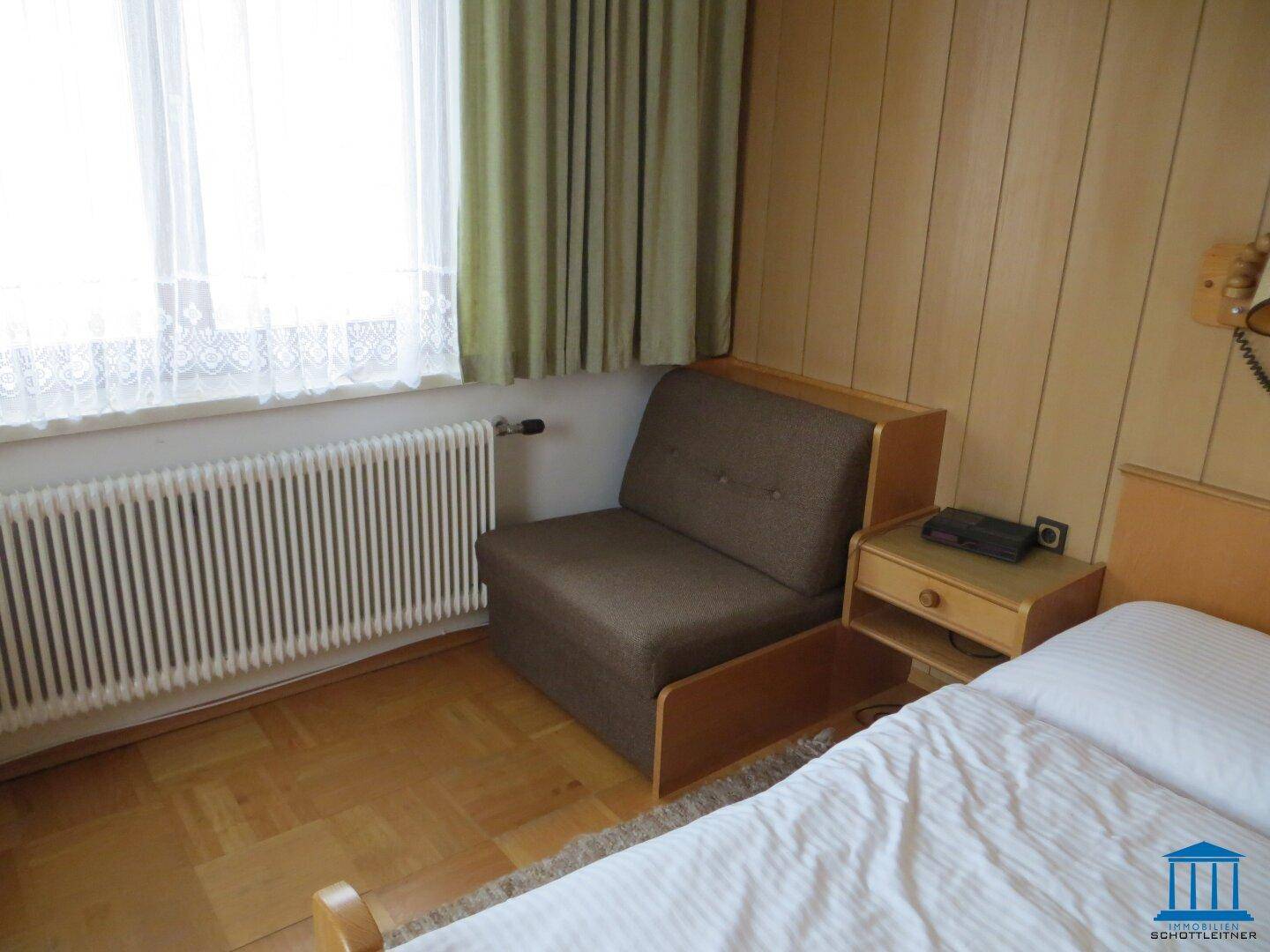 Schlafzimmer mit Einzelbett, Sessel, Heizkörper und Holzvertäfelung an der Wand.