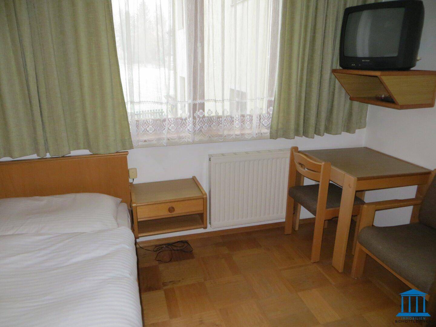 Schlafzimmer mit Bett, kleinem Tisch, Stühlen und einem Fernseher an der Wand.